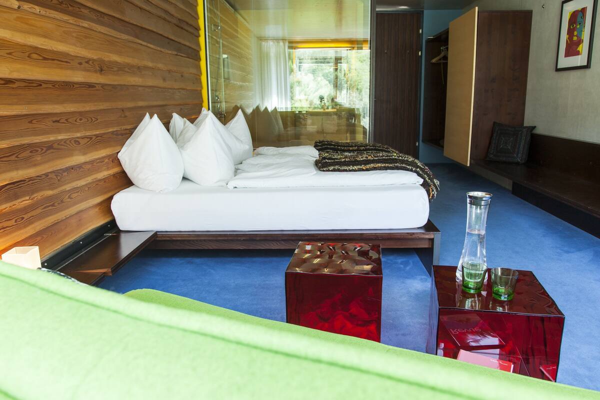 A modern room with a wooden wall, a bed with white bedding, a red cube-shaped object, and a bench with a cushion.