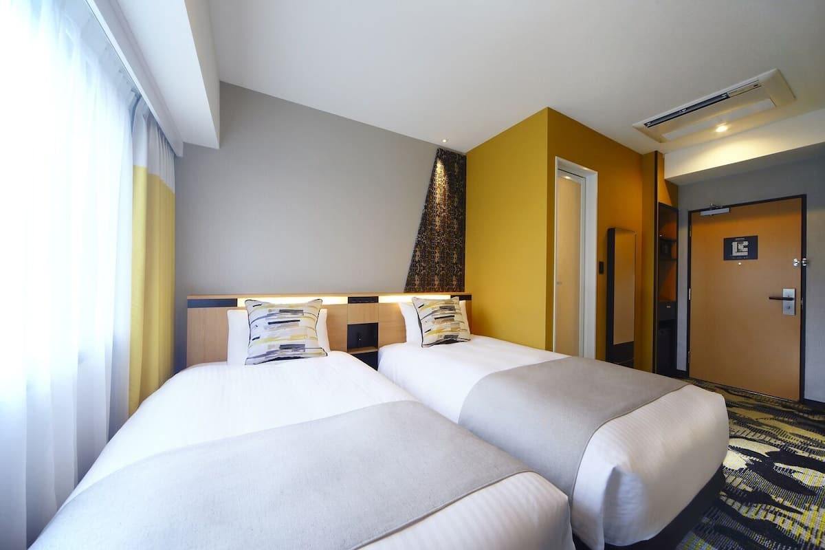 Habitación de hotel con dos camas, cabecero, lámpara de pared y una puerta.