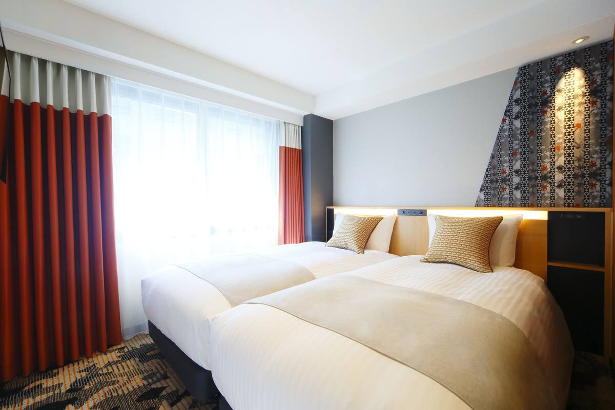 Habitación de hotel con dos camas, un ventanal con cortinas, una pared con diseño y una mesita de noche con lámpara.