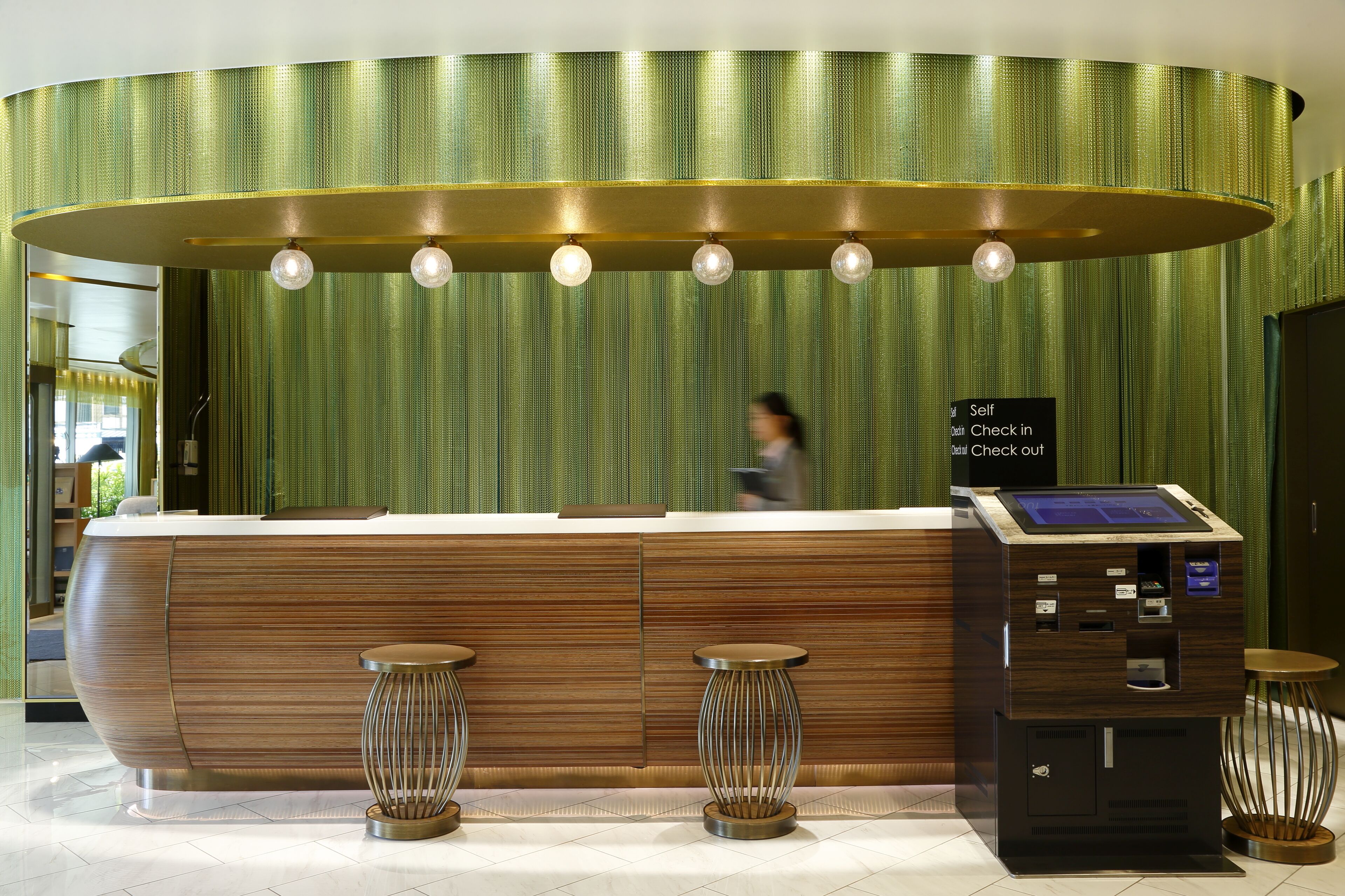 A reception desk with a self-check-in kiosk, a green textured wall, and a person walking in the background.