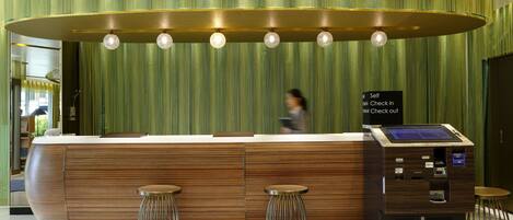 A reception desk with a self-check-in kiosk, a green textured wall, and a person walking in the background.