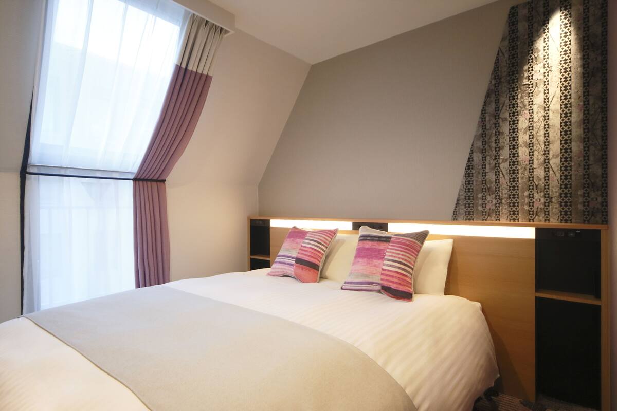 Una cama bien hecha con cabecera, una ventana con cortinas traslúcidas y una cortina estampada a la derecha.