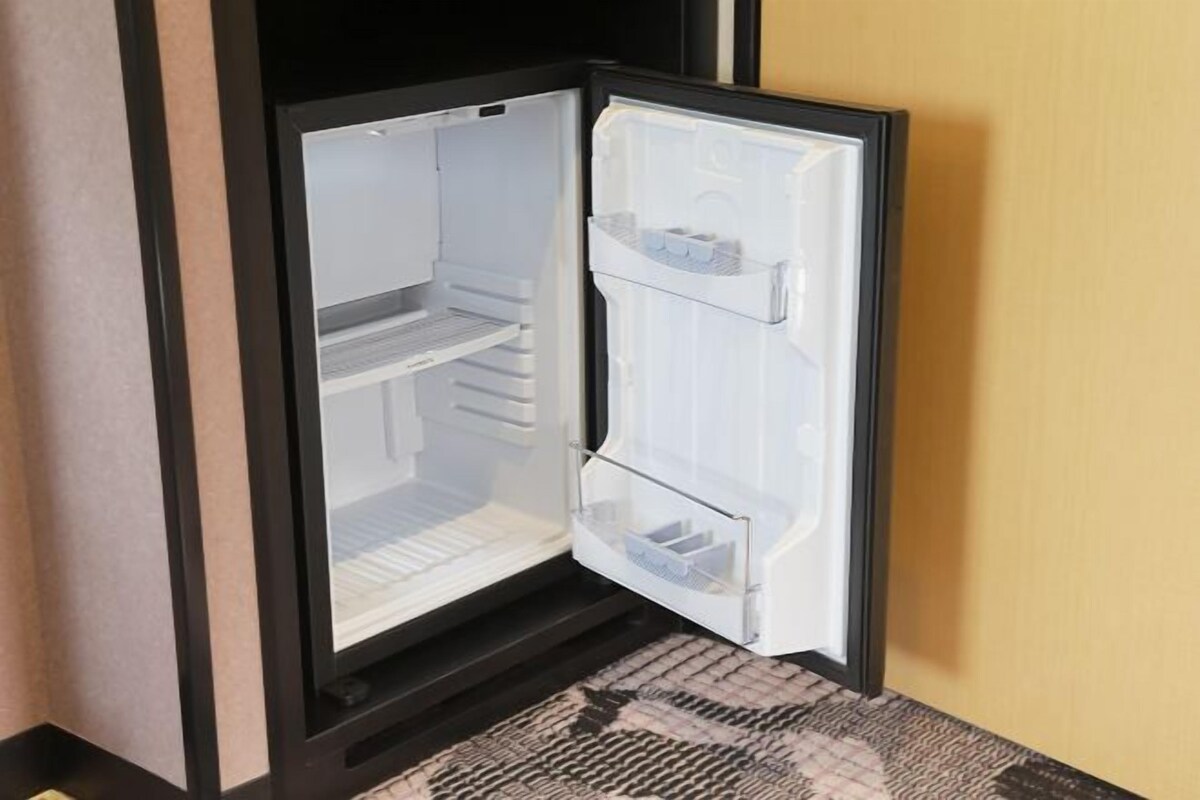 A modern, empty refrigerator with a clear door, located in a room with a patterned carpet and a plain wall.