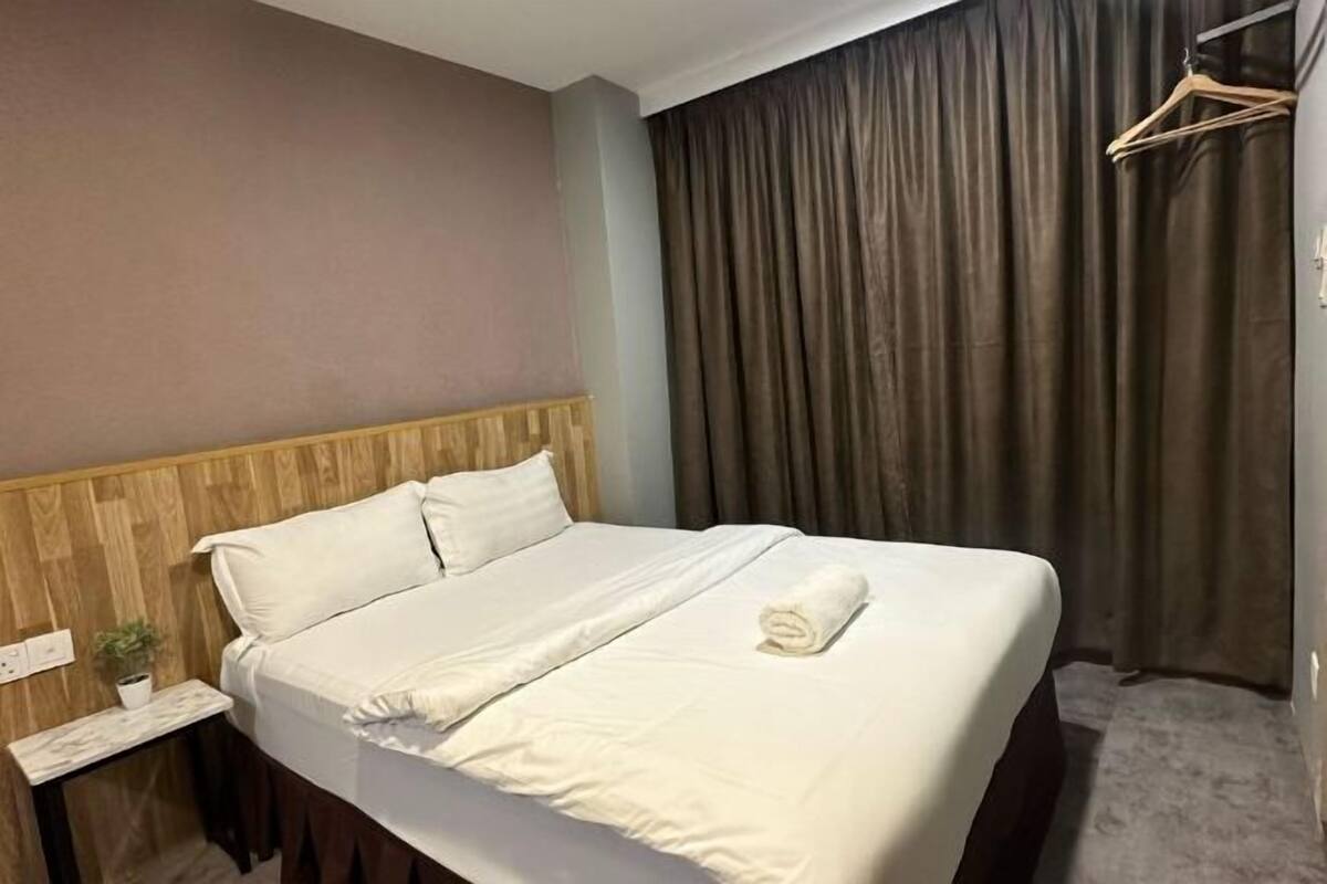 A neatly made bed with white linens, a wooden headboard, a small bedside table with a plant, and a dark curtain covering a window.