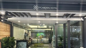 Exterior del Hotel D'Garden con una mesa de recepción, plantas en macetas y una entrada de cristal.