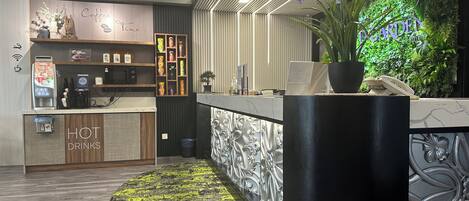 Una zona de recepción moderna con estación de café, un letrero que dice 'HOT DRINKS' y una pared verde con la palabra 'GARDEN'.