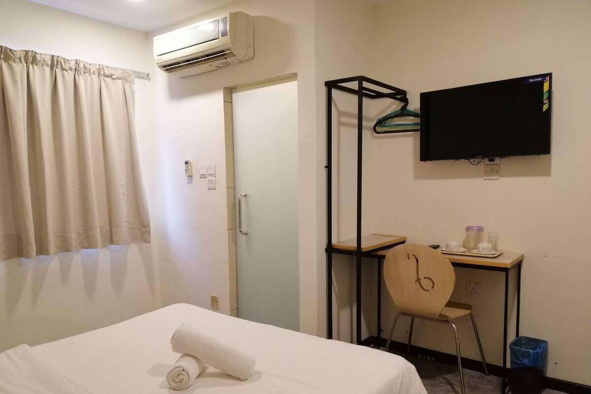 A hotel room with a bed, a desk, a chair, a TV, and an air conditioning unit.