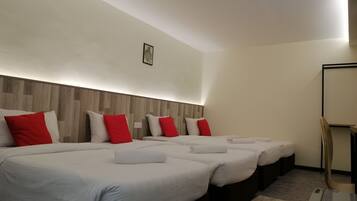 Quarto de hotel com quatro camas dispostas em linha, cada uma com roupa de cama branca e almofadas vermelhas. Possui cabeceira de madeira, um pequeno quadro na parede e um espelho no lado direito.
