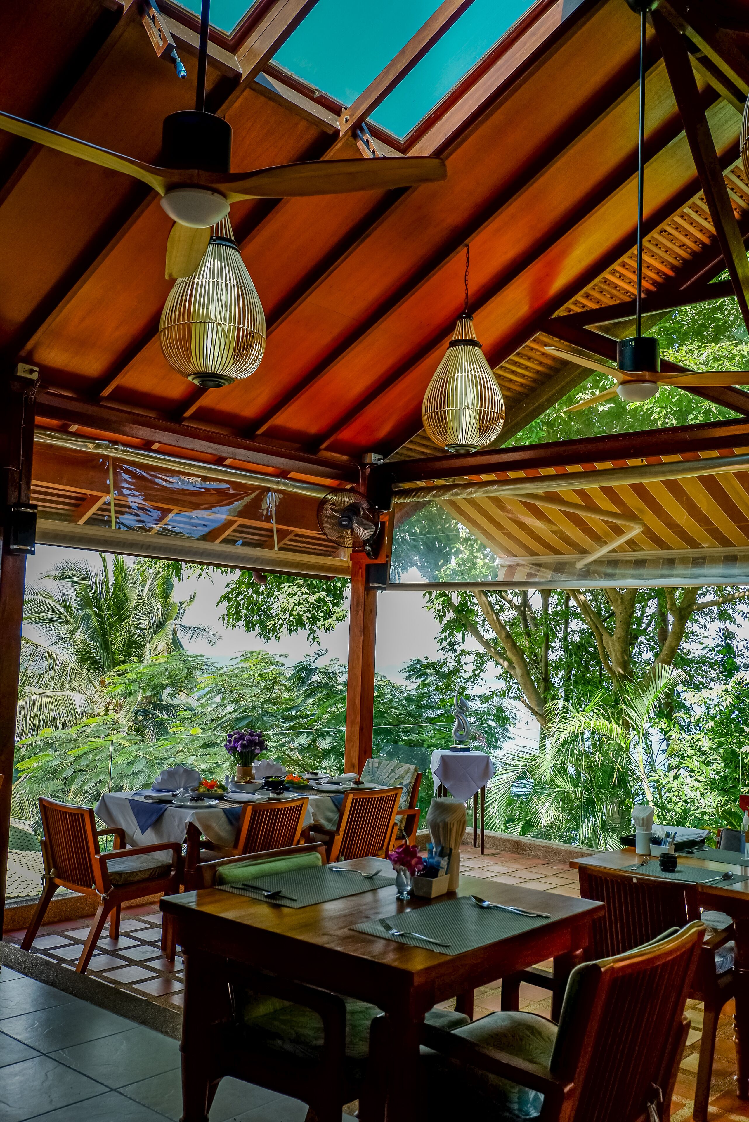 A covered outdoor dining area with wooden tables and chairs, hanging light fixtures, and a view of lush greenery.
