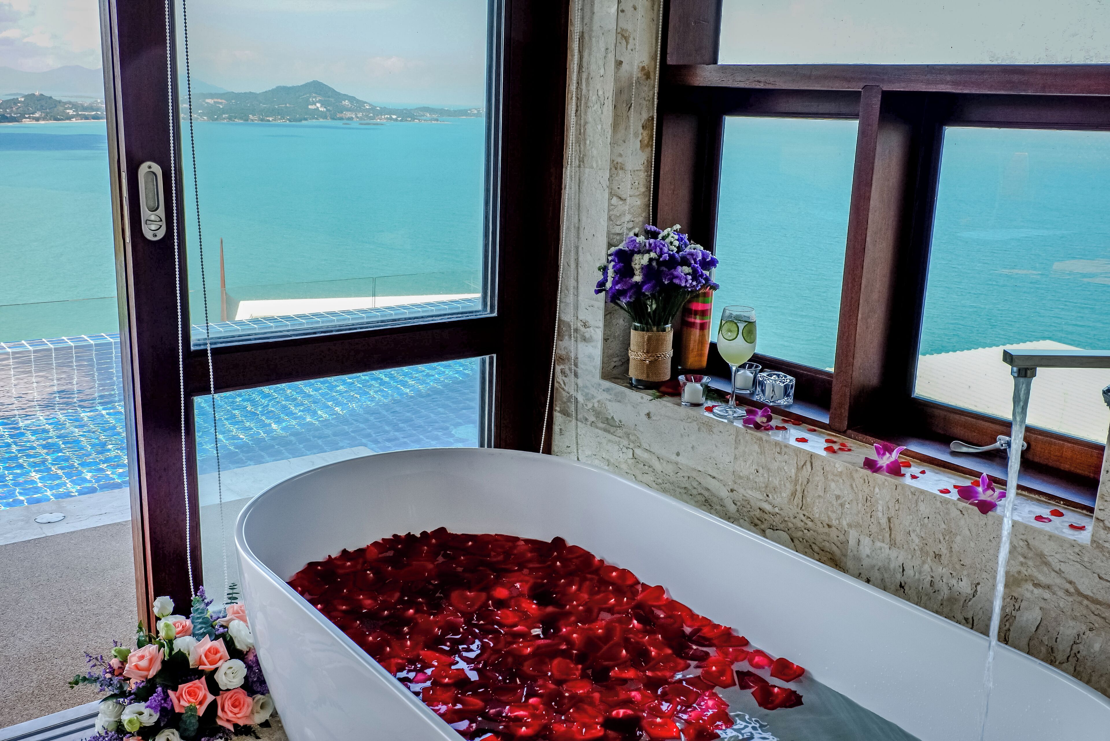 A bathtub filled with rose petals and water, a window with a view of the sea, and a vase with flowers on the windowsill.