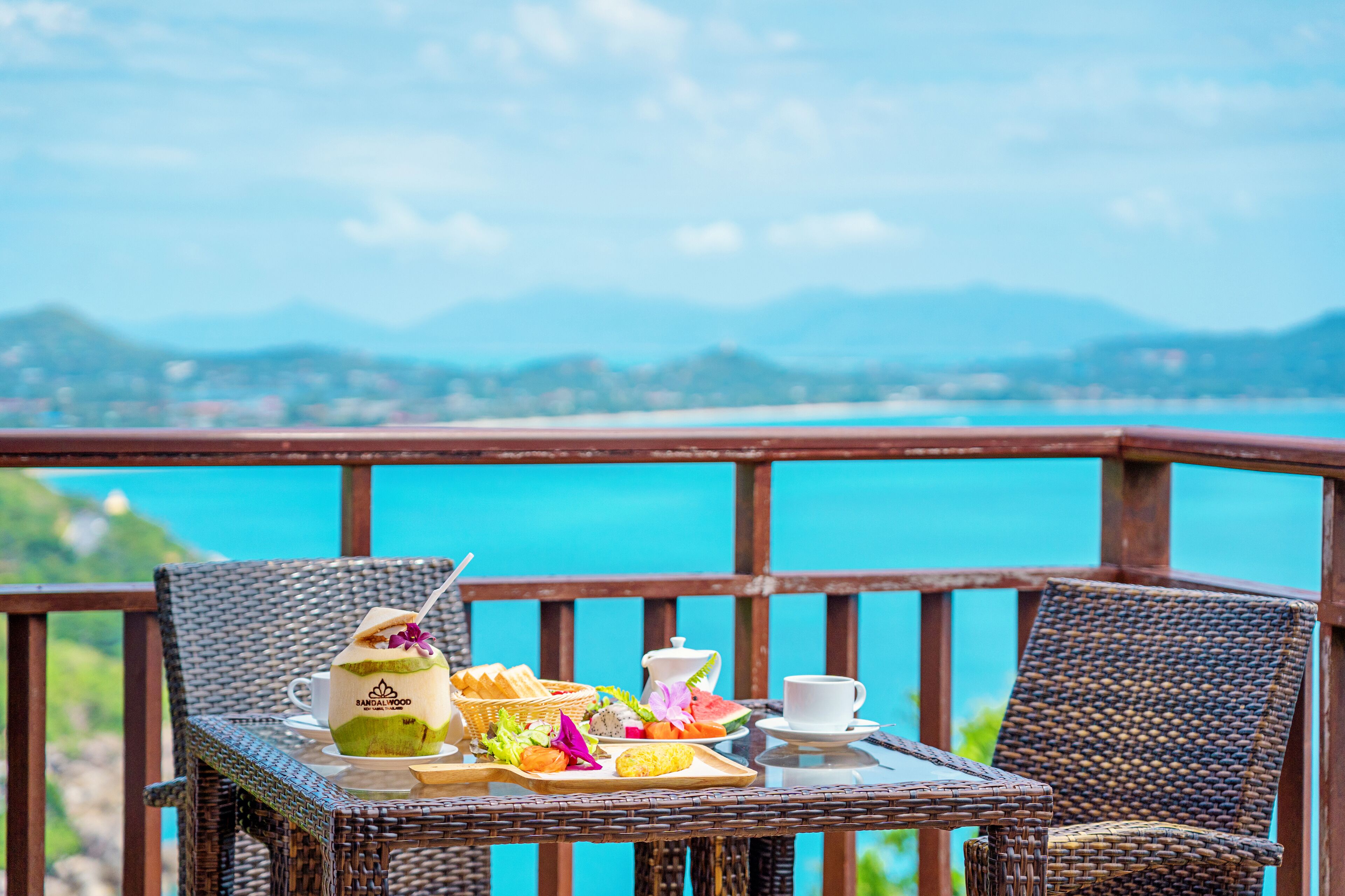 A wicker table with a breakfast tray, a coconut drink, and a cup on a balcony overlooking a turquoise sea and distant mountains.