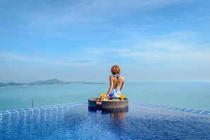 Une personne est assise sur une plateforme flottante, avec un panier contenant des boissons et un verre de jus d’orange, et elle a devant elle une étendue d’eau et, au loin, une terre ferme.
