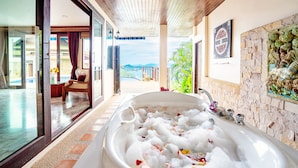Een luxe badkamer met een ruime badkuip vol bubbels en rozenblaadjes, een glazen douchecabine en uitzicht op een zwembad en bergen.