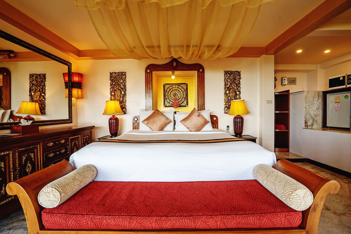 Een groot bed met een rode sprei en twee houten hoofdborden, een grote spiegel, en een muur met een decoratief wandtapijt.