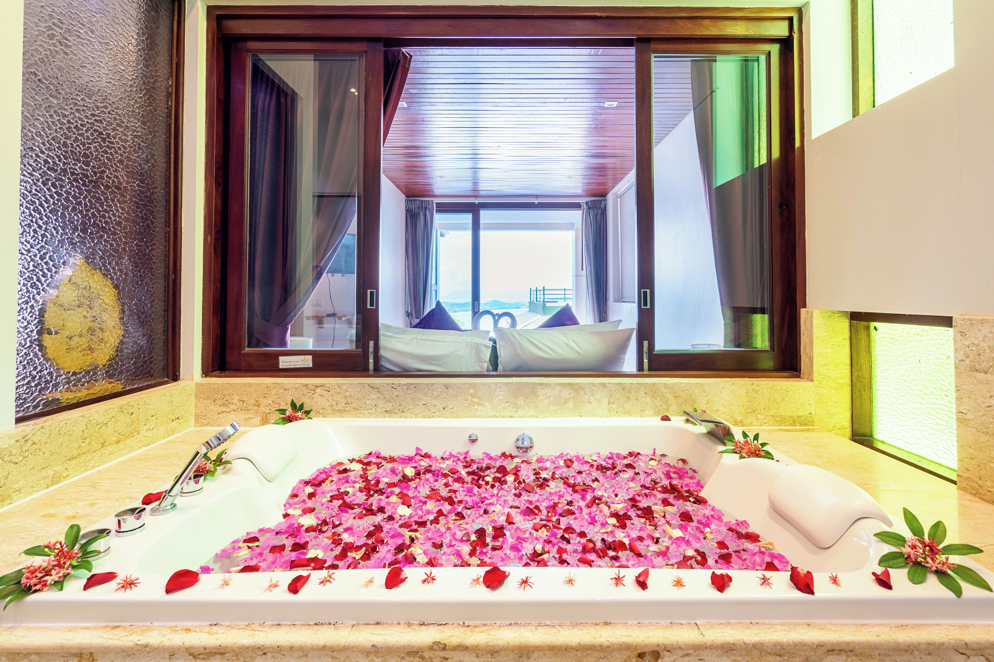 A bathroom with a bathtub filled with rose petals, a large window, and a view of a bedroom with a bed and curtains.