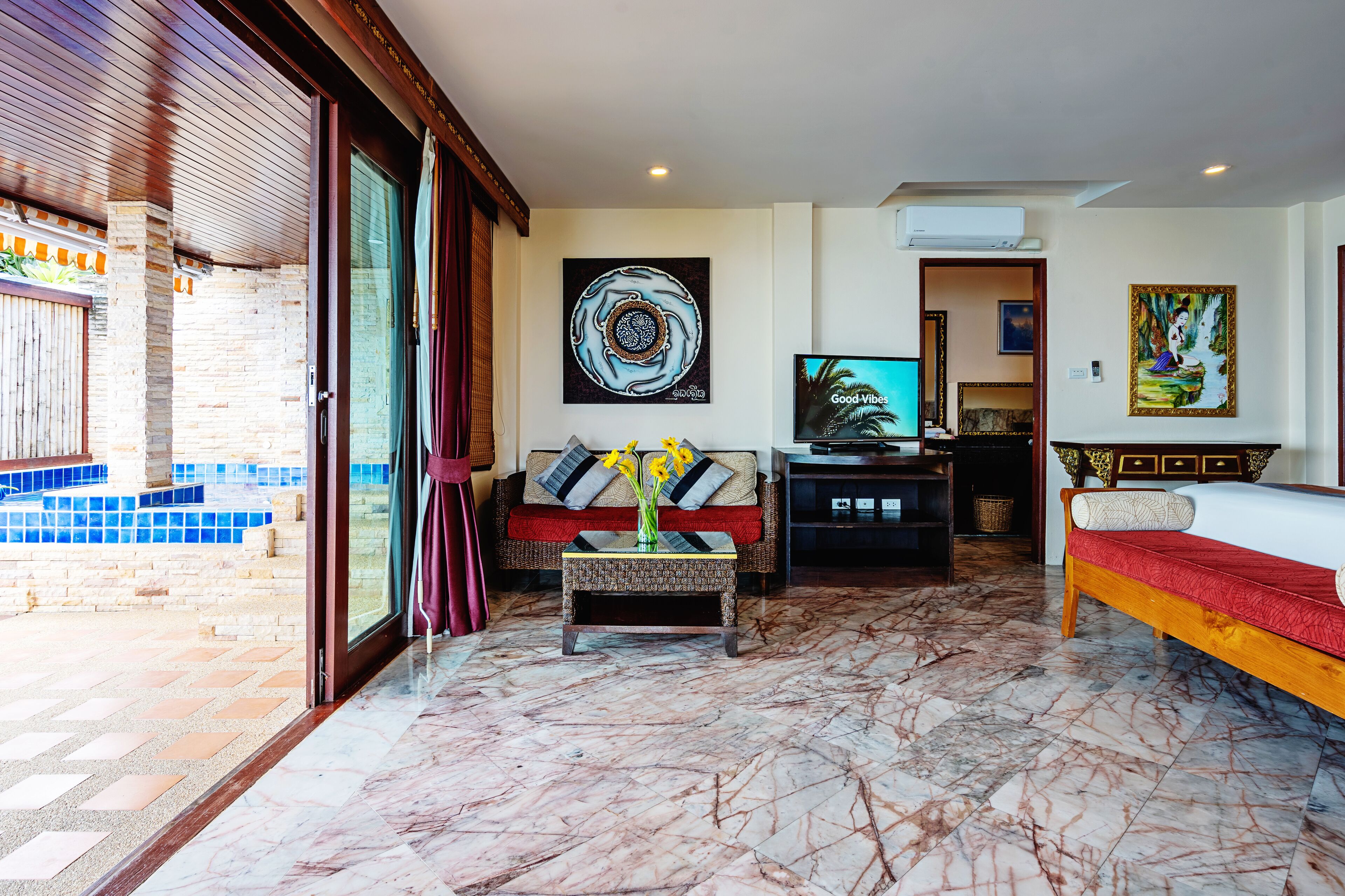 A spacious living area with a large TV, a sofa, a coffee table with flowers, and a bed with a red bedspread.