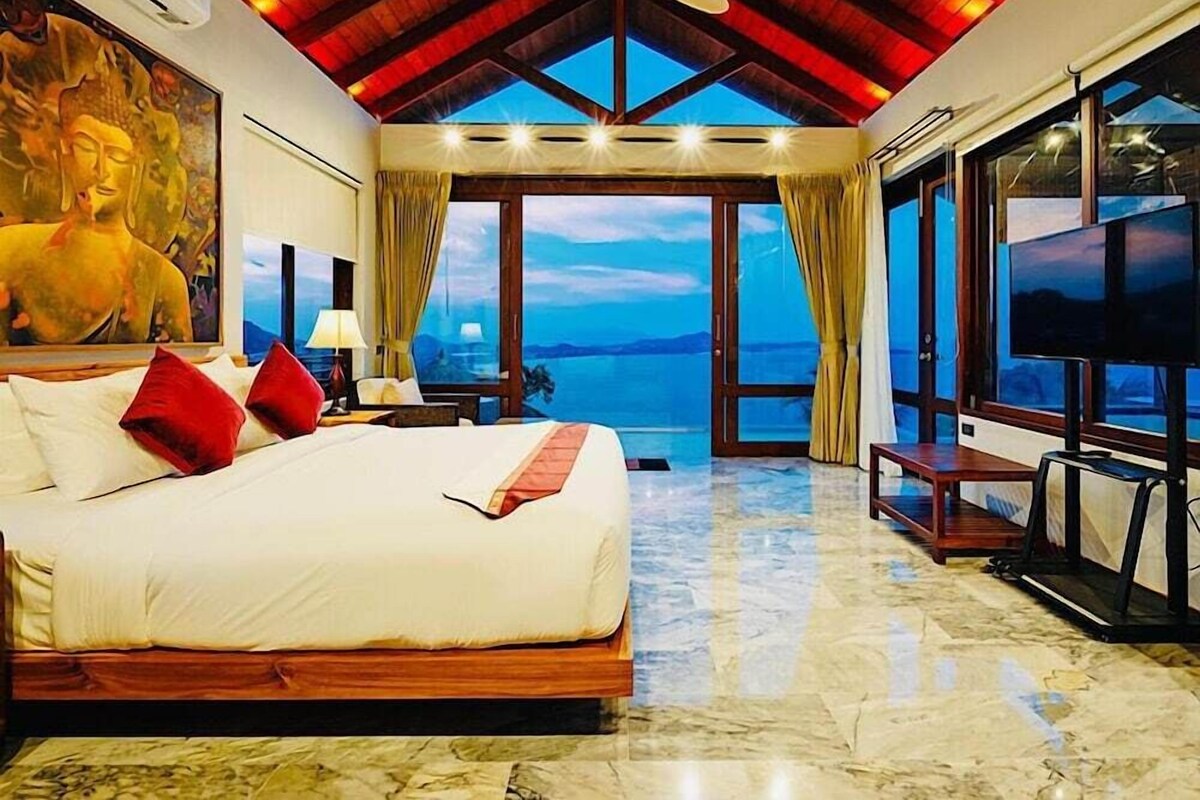 Une chambre d’hôtel avec un grand lit, un ventilateur de plafond, une statue de Bouddha, une télévision et une vue sur la mer.
