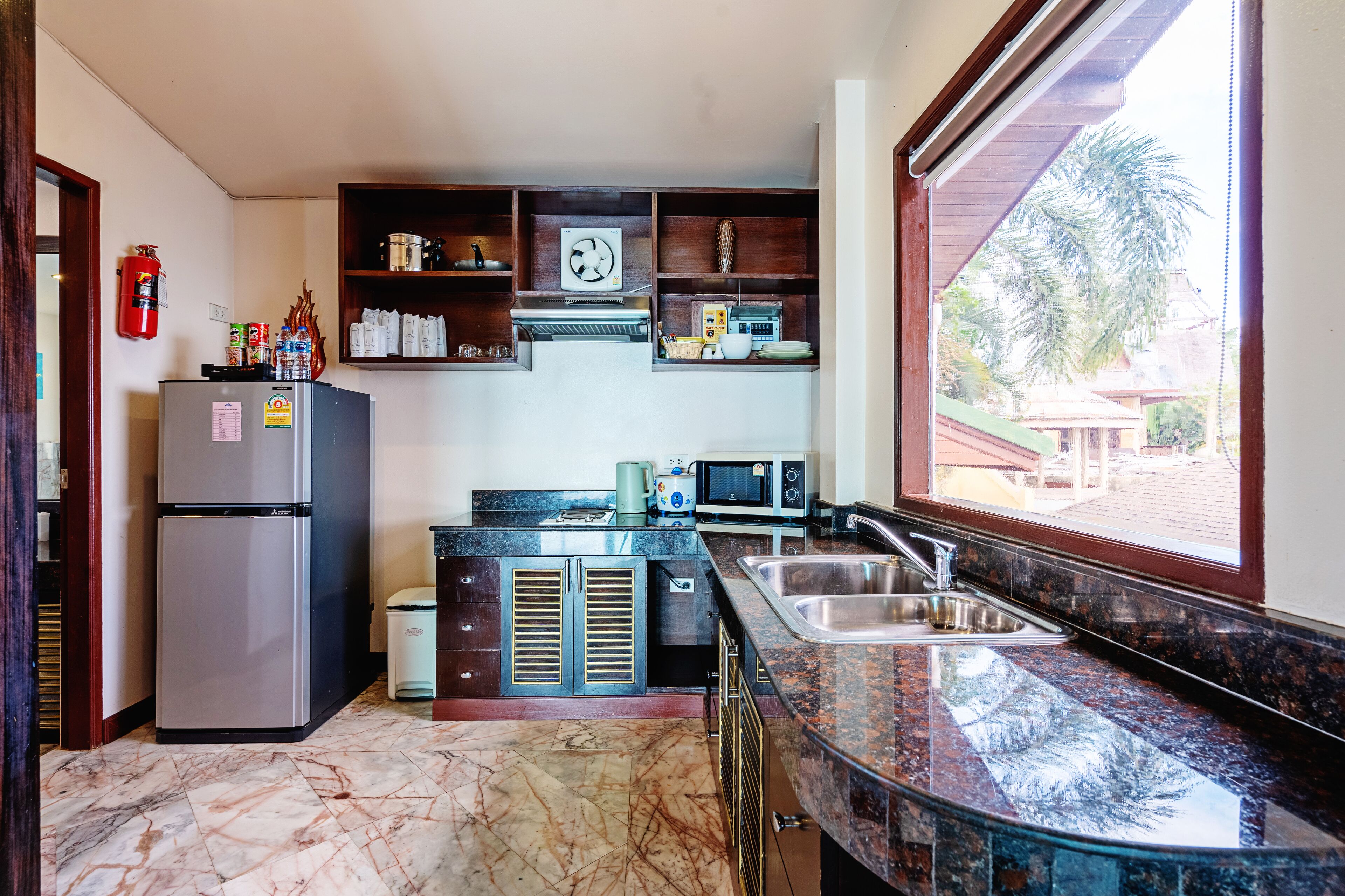 A compact kitchen with a refrigerator, microwave, and sink. Shelves above the counter hold various kitchen items.