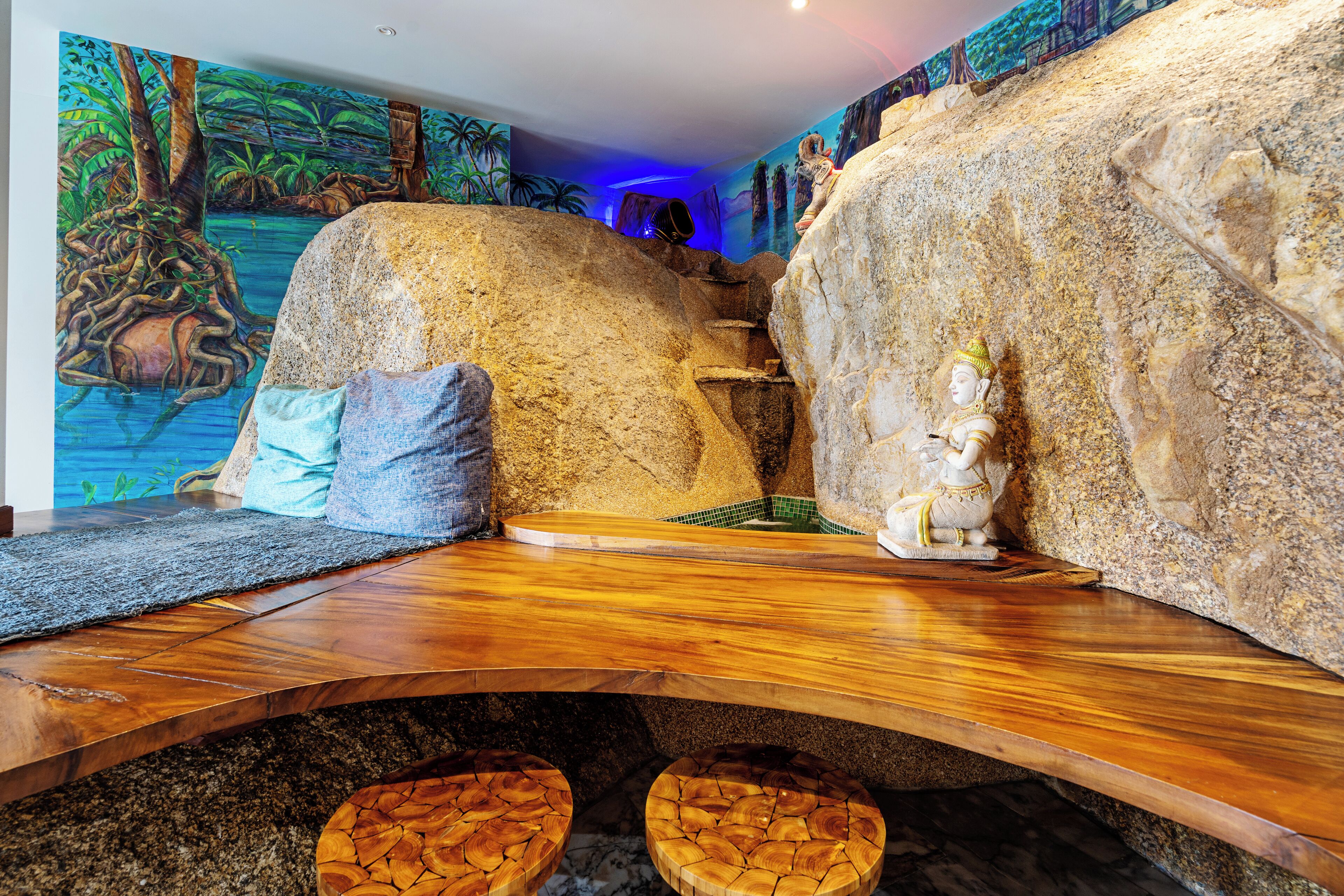 Una habitación con una mesa de madera, dos taburetes, un mural de una escena selvática y una estatua sobre una roca.