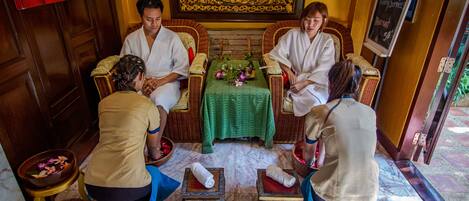 A traditional spa setting with four people receiving a massage. There are two massage tables, a small table with flowers, and a decorative wall.