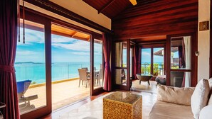 Een ruime kamer met een grote schuifpui naar een balkon met uitzicht op de oceaan. Er staat een houten salontafel en een beige bank met kussens.