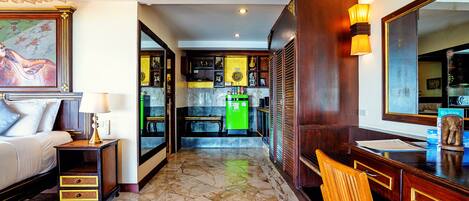 Habitación de hotel con cama, mesita de noche, lámpara y escritorio con silla. Se observa una zona de cocina al fondo con refrigerador y armarios.