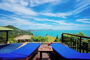 Zona junto a la piscina con sillas reclinables azules, una mesa de madera y vistas al mar y montañas a lo lejos.