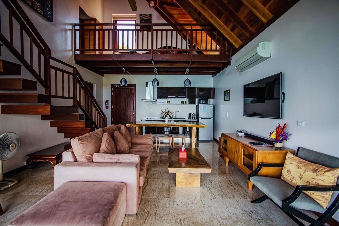 A spacious living area with a high ceiling, a wooden staircase, a kitchen with modern appliances, and a flat-screen TV mounted on the wall.