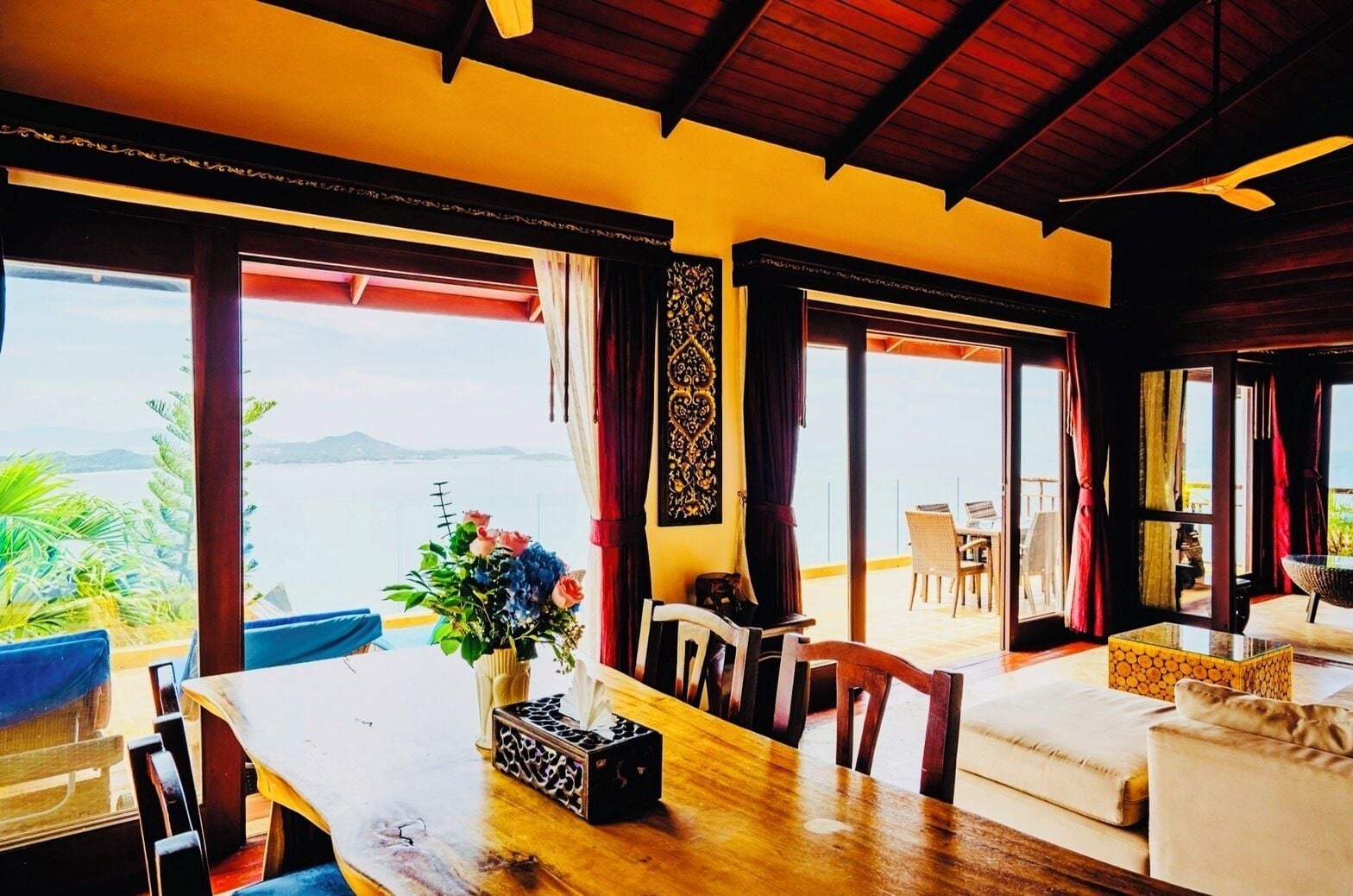 A dining area with a wooden table, chairs, and a vase of flowers. There are large windows offering a view of the sea and mountains.