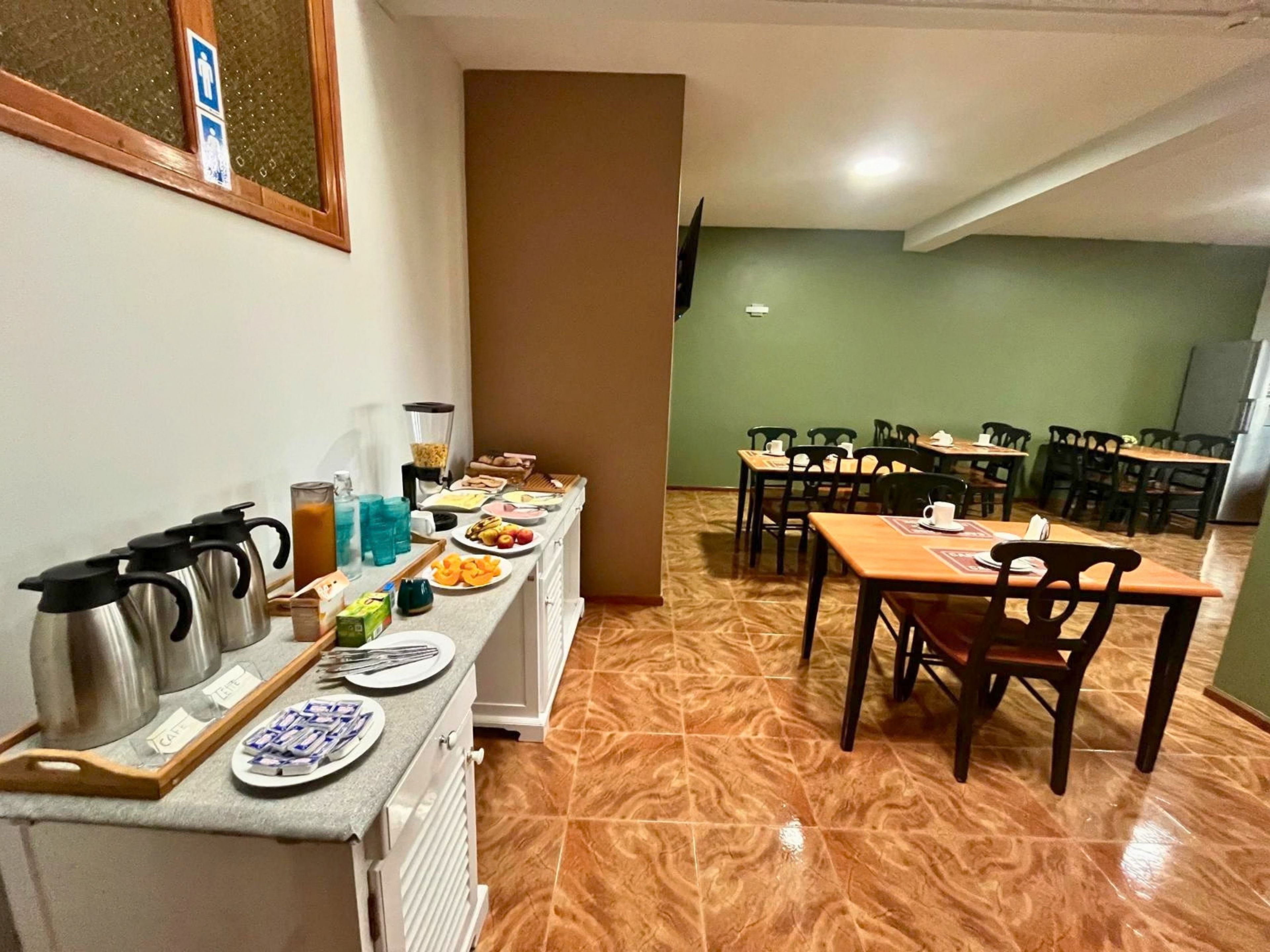 Uma área de refeições com várias mesas e cadeiras, um balcão de buffet com alimentos e um espaço para café com chaleiras e xícaras.