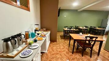 Uma área de refeições com várias mesas e cadeiras, um balcão de buffet com alimentos e um espaço para café com chaleiras e xícaras.