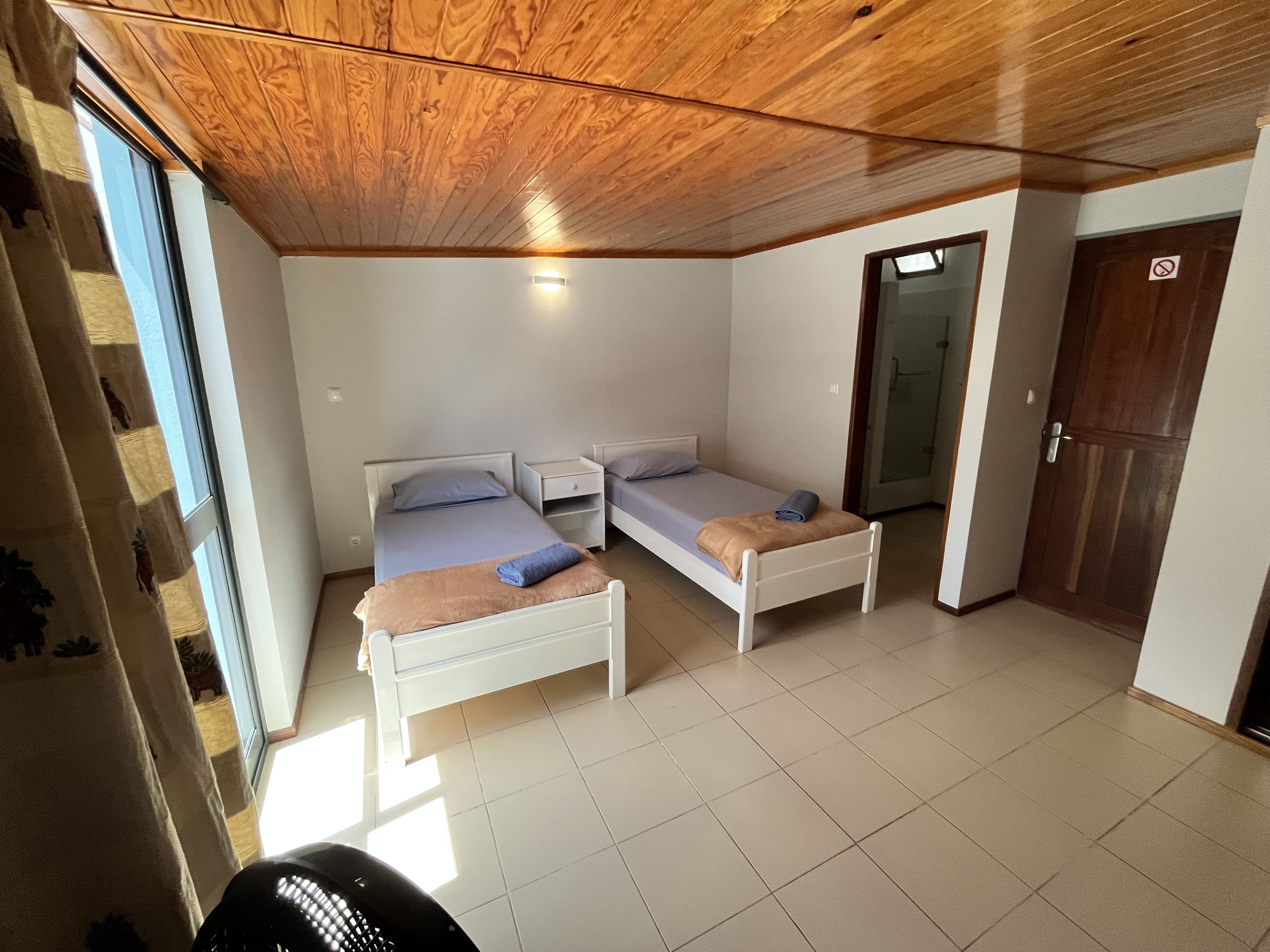 Quarto com duas camas de solteiro, teto de madeira, porta branca e uma janela.