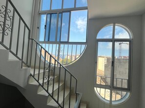 A staircase with a metal railing leading to a landing with large windows offering views of buildings and a crane.