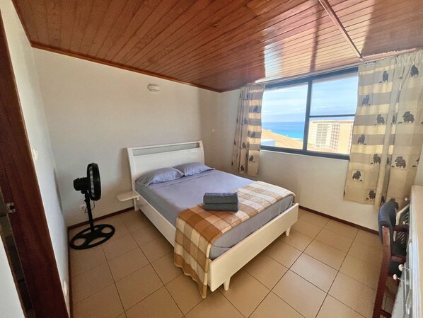 A bedroom with a bed, a standing fan, a window with curtains, and a view of the beach.