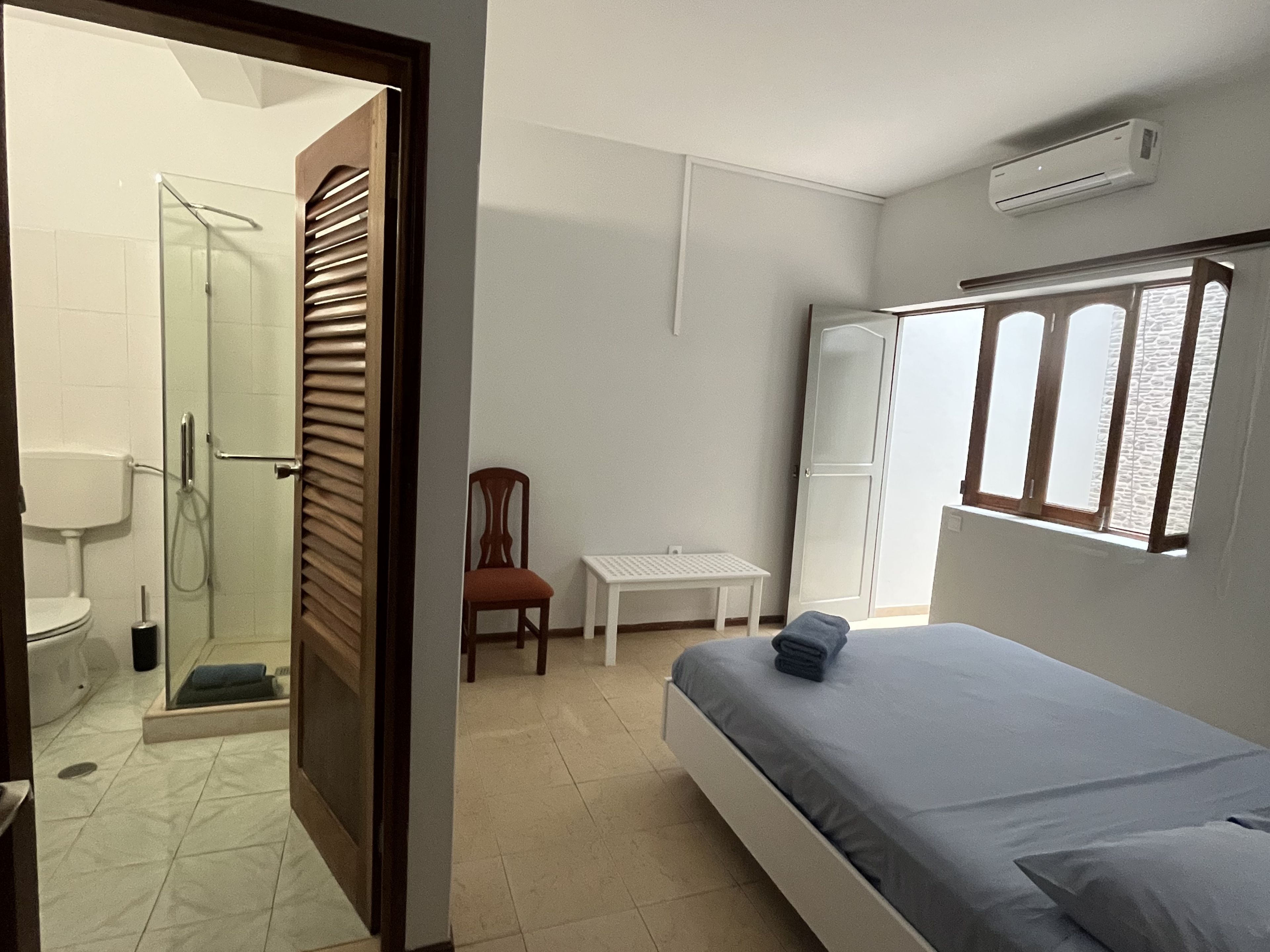 Un dormitorio con cama, una silla, una mesita, un armario y un baño con ducha.