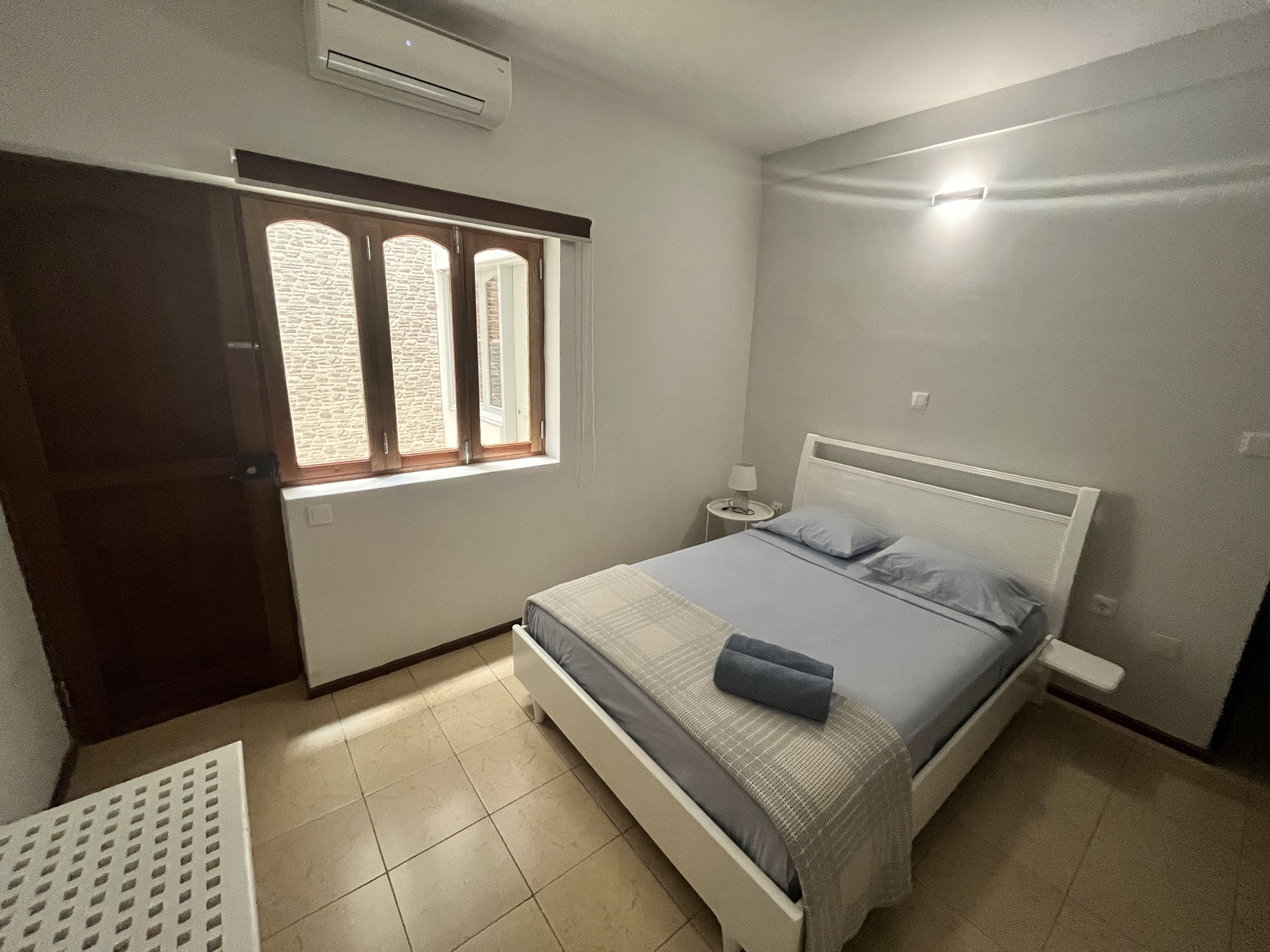 A bedroom with a bed, a nightstand, a lamp, a window with curtains, and an air conditioning unit.