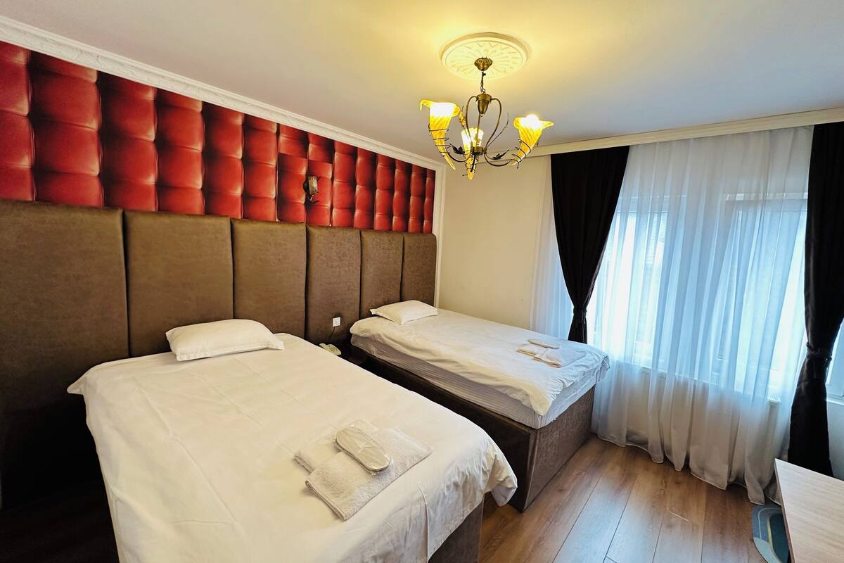 Habitación de hotel con dos camas, un candelabro, un cabecero acolchado rojo y un ventanal con cortinas traslúcidas.