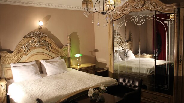 A hotel room with a large bed, a nightstand, a lamp, a mirror, and a black leather chair. - Hotel Adria (Pristina)