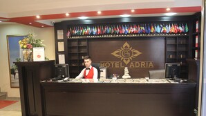 A hotel reception desk with a receptionist, a display of flags, and a logo for HOTEL ADRIA. - Hotel Adria (Pristina)