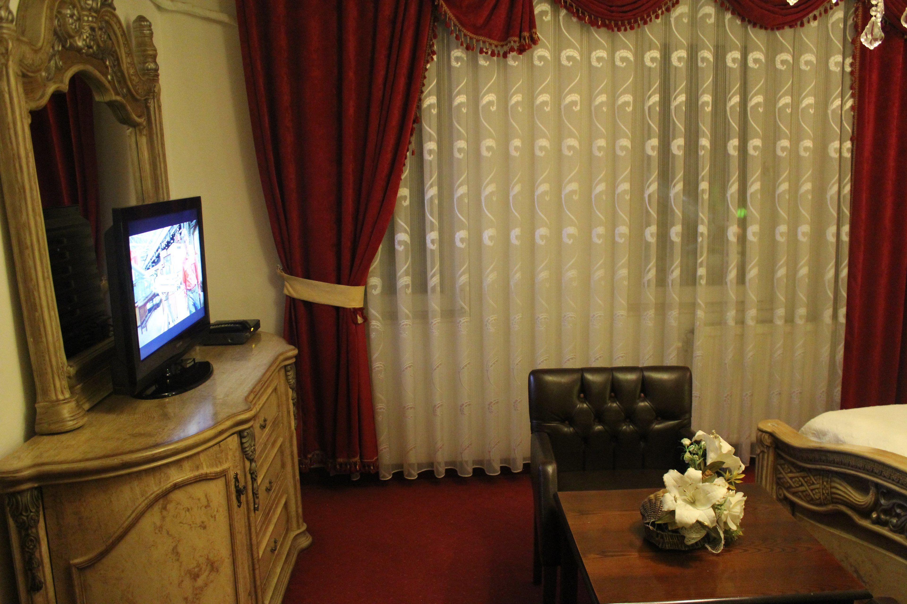 Une chambre d’hôtel avec un téléviseur posé sur un meuble en bois, un fauteuil en cuir, une table en bois ornée d’un vase de fleurs et un lit recouvert d’une couette blanche.