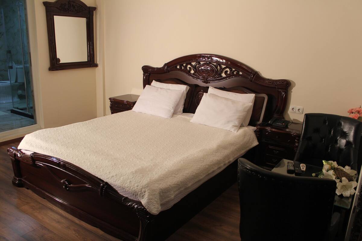 Una cama bien hecha con una colcha blanca y dos almohadas, una mesita de noche de madera con un teléfono, y una silla con un florero de flores.
