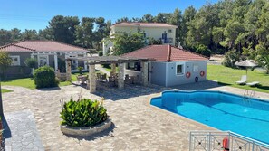 A pool area with a stone patio, a covered seating area, and a small building with a red-tiled roof.