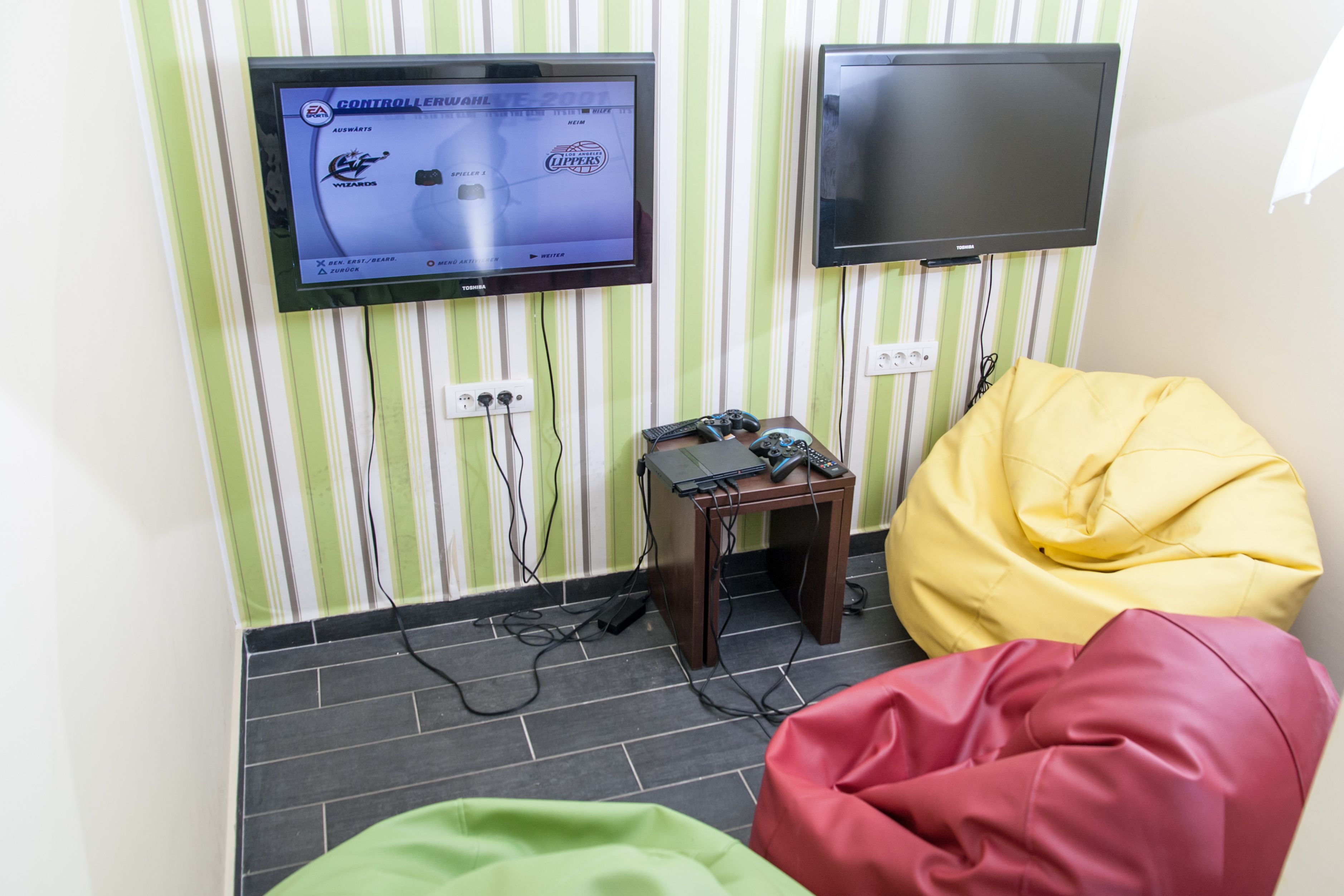 Una habitación con dos televisores montados en la pared, una mesita con controles de videojuegos y puffs en verde, amarillo y rosa.