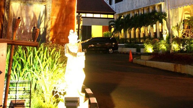 A traditional pagoda-style structure with a statue, adjacent to a modern multi-story building with balconies and a parking area. - Puri Denpasar Hotel (Jakarta)