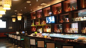 A bar area with a long counter, multiple bar stools, shelves stocked with bottles, and a television mounted on the wall. - Puri Denpasar Hotel (Jakarta)