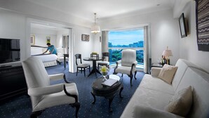 A hotel room with a bed, a TV, a dining area with chairs and a table, a sofa, and a view of the city through large windows. - Puri Denpasar Hotel (Jakarta)