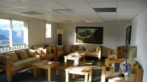 A spacious living area with multiple seating options, including sofas and chairs, arranged around wooden tables. There is a large window providing a view of mountains, and a painting is displayed on the wall. - Les Adrets de Peyragudes (Germ)
