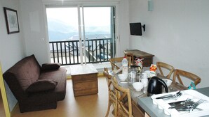 A living room with a couch, a dining table set with cutlery, and a view of mountains through a balcony door. - Les Adrets de Peyragudes (Germ)