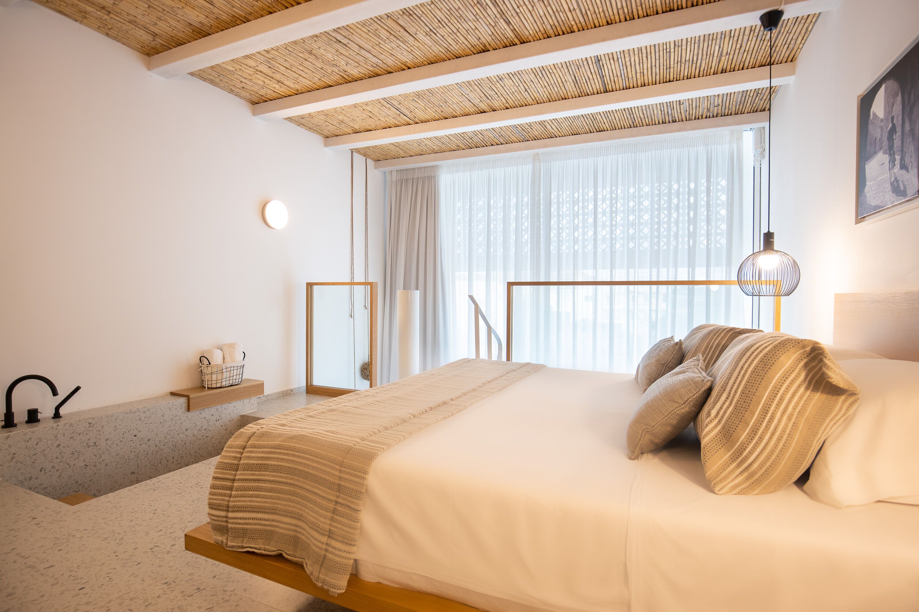 A modern bedroom with a bed, a bedside table, a window with sheer curtains, a hanging light fixture, and a wooden ceiling.