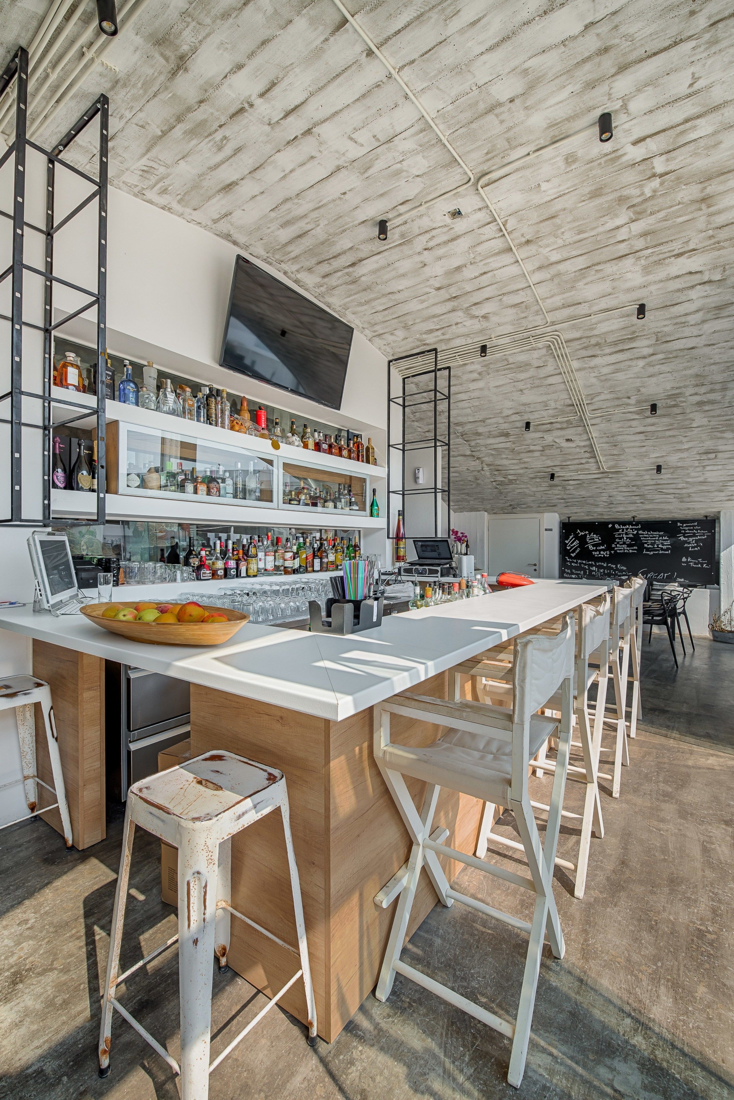 A modern bar area with a long counter, white stools, and shelves stocked with bottles.